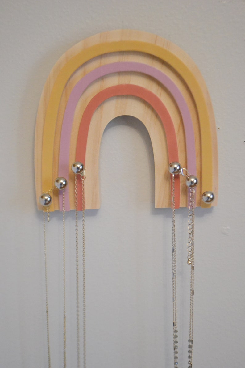 May include: Wooden rainbow-shaped jewelry organizer with a natural wood finish and colorful arches in yellow, pink, and coral. Silver-colored ball-shaped knobs hold multiple necklaces of varying lengths. A decorative and functional item.