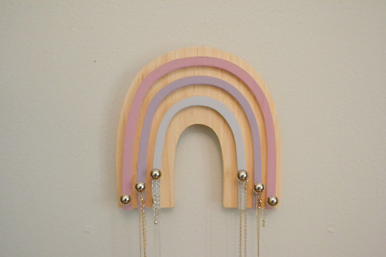 May include: A wooden rainbow-shaped jewelry holder with three layers of pastel pink, lavender, and gray paint. The holder has gold-colored metal hooks and beads.