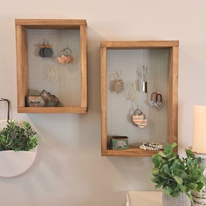 May include: Two rectangular wooden wall-mounted jewellery organisers with wire mesh backing. Each frame displays various hanging earrings and small accessories on the mesh, with additional items resting on the bottom wooden ledge of each frame.