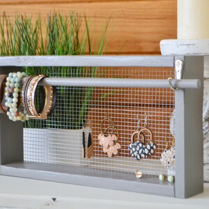 May include: A gray wooden jewelry organizer with a wire mesh backing. The organizer displays bracelets and earrings. The bracelets are on a horizontal bar, and the earrings hang from the mesh. The background includes candles and greenery.