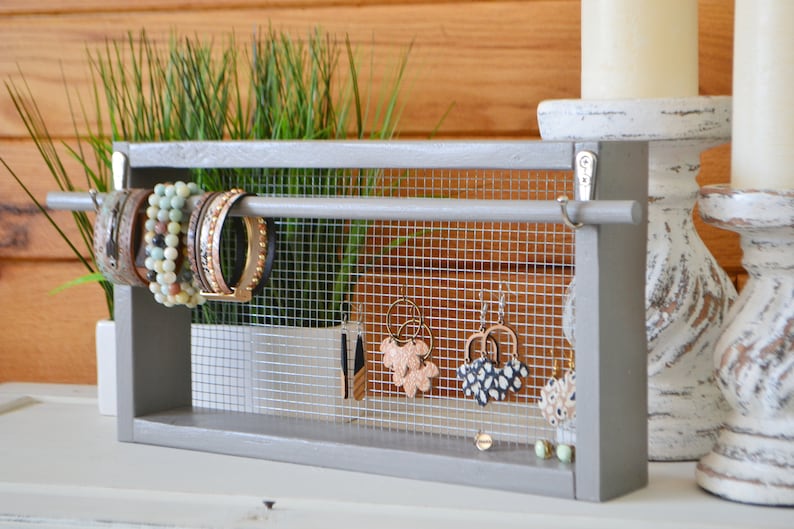 May include: A gray wooden jewelry organizer with a wire mesh backing. The organizer displays bracelets and earrings. The bracelets are on a horizontal bar, and the earrings hang from the mesh. The background includes candles and greenery.