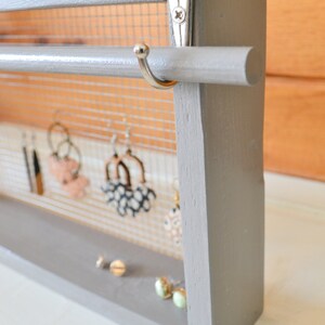 May include: A gray wooden earring holder with a mesh backing and a dowel rod for hanging earrings. Several pairs of earrings are displayed, along with a few pairs of stud earrings on the base. The holder is painted gray and has a simple, modern design.