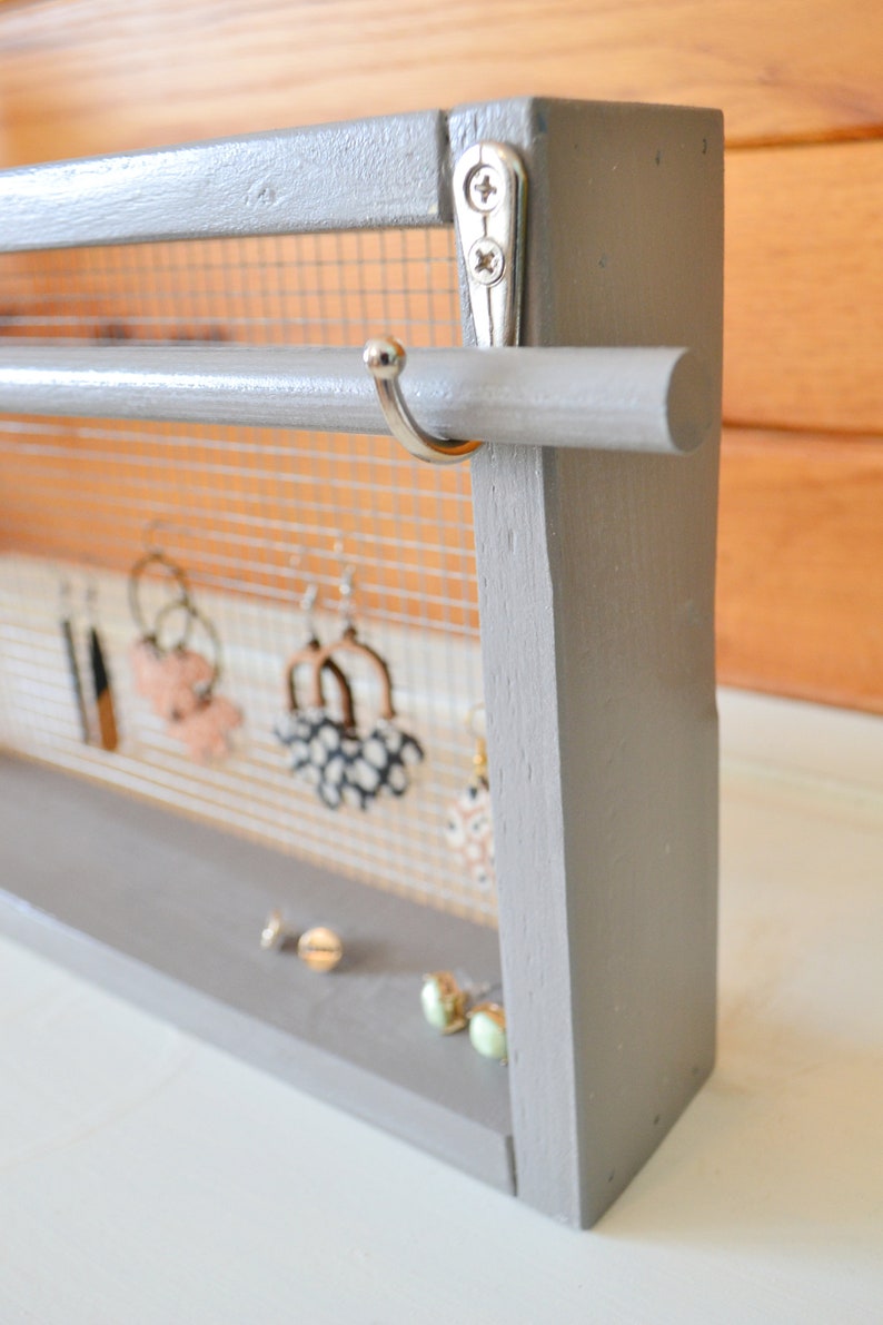 May include: A gray wooden earring holder with a mesh backing and a dowel rod for hanging earrings. Several pairs of earrings are displayed, along with a few pairs of stud earrings on the base. The holder is painted gray and has a simple, modern design.