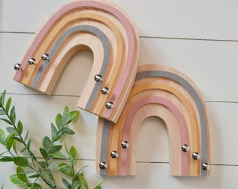 May include: Two wooden rainbow key holders with a pink, yellow, and gray color scheme. Each rainbow has five metal hooks.