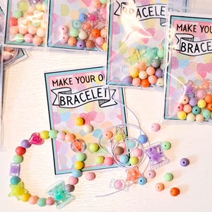 May include: A DIY bracelet kit with colorful beads in various shapes and sizes. The kit includes clear plastic bags with beads, a completed bracelet, and cards that read "MAKE YOUR OWN BRACELET". The beads are in shades of pink, yellow, orange, green, and purple.