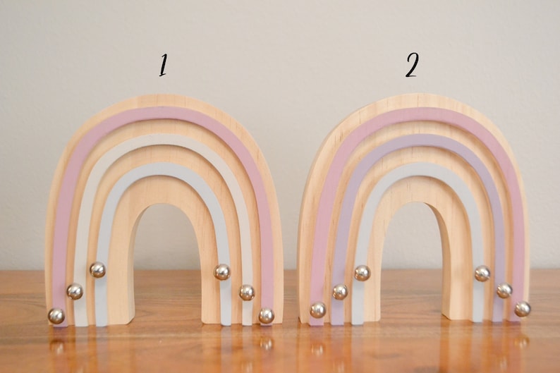 May include: Two wooden rainbow wall decor pieces with a pastel pink, light purple, and white color scheme. Each rainbow has five silver metal balls hanging from the bottom.