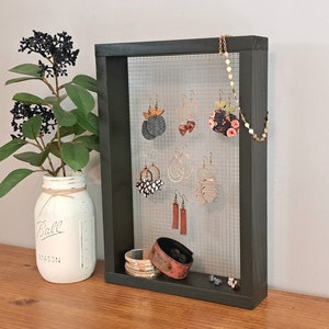 May include: Black framed jewelry organizer with a wire mesh backing. The organizer has a variety of earrings and a necklace hanging on it. There is a brown leather cuff bracelet and a white mason jar with a black label that says "Ball Mason" on a wooden surface.