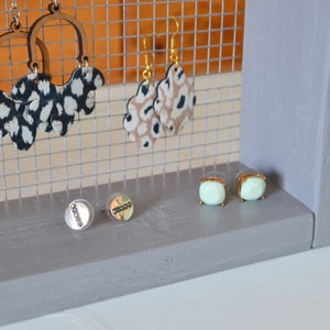 May include: A gray wooden earring display stand with a wire mesh backing. The stand holds various earrings, including wooden hoops with black and white patterned accents, tan and black patterned earrings, and mint green square stud earrings.