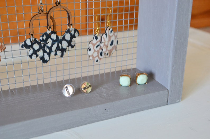 May include: A gray wooden earring display stand with a wire mesh backing. The stand holds various earrings, including wooden hoops with black and white patterned accents, tan and black patterned earrings, and mint green square stud earrings.