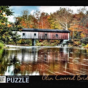 May include: A red, white, and gray covered bridge spans a river with a reflection of the bridge and surrounding trees in the water. The bridge is surrounded by fall foliage in shades of red, orange, yellow, and green. The text "Olin Covered Bridge" is written in yellow cursive script below the image.
