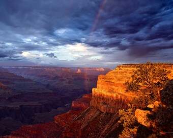 May include: A vibrant rainbow arches over the Grand Canyon, with dramatic clouds casting shadows on the red rock formations. The sun illuminates the canyon walls, creating a warm glow.
