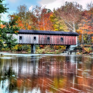 May include: A wooden covered bridge with a red and white painted exterior, spanning a river with a reflection of the surrounding autumn foliage. The bridge has three visible windows on each side.