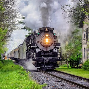 May include: A vintage black steam train with the number 765 on the front, billowing white smoke as it travels along a railroad track. The train is surrounded by lush green trees and grass, with a house visible in the background.