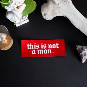 Puede incluir: Parche de tela rojo con texto blanco que dice "this is not a man." El parche está sobre una superficie negra con un hueso blanco, una pequeña cúpula de vidrio con un objeto blanco en su interior y un cristal morado.