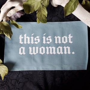 May include: A light blue fabric banner with the text "this is not a woman." printed in white.