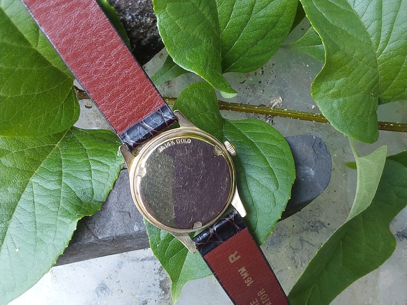 May include: A gold-tone wristwatch with a brown leather band. The back of the watch case is visible and says "14K Gold".