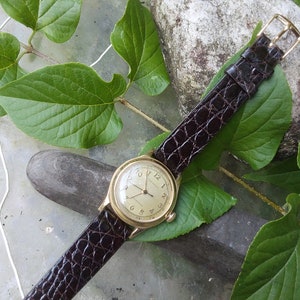 May include: A gold wristwatch with a white face and black Roman numeral hour markers. The watch has a brown leather band with a crocodile pattern. The watch is lying on a gray surface with green leaves.