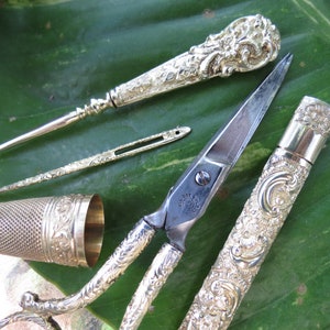 May include: A set of antique silver sewing tools, including a thimble, a needle threader, a pair of scissors, and a needle case. The tools are decorated with intricate floral patterns.