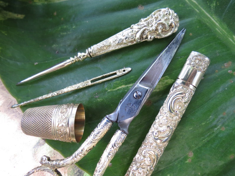 May include: A set of antique silver sewing tools, including a thimble, a needle threader, a pair of scissors, and a needle case. The tools are decorated with intricate floral patterns.