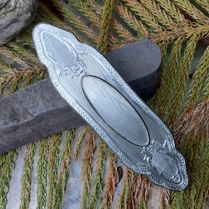 May include: A silver-colored metal bookmark with an ornate design. The bookmark has a rectangular shape with a rounded top and bottom. The design features a central oval shape with a textured surface and intricate floral patterns around the edges.