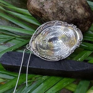 May include: Silver tea strainer with a decorative floral design and a long handle. The strainer is sitting on a black surface.