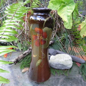 May include: A brown ceramic vase with a floral design. The vase has a wide mouth and a narrow base. The floral design features a large red flower with yellow accents and green leaves.