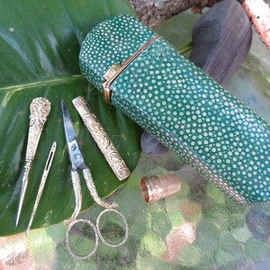 May include: A green leather sewing kit with a white polka dot pattern. The kit includes a pair of scissors, a needle threader, a thimble, and a small case with a needle inside. The kit is lying on a green leaf.
