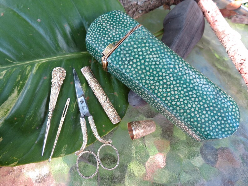 May include: A green leather sewing kit with a white polka dot pattern. The kit includes a pair of scissors, a needle threader, a thimble, and a small case with a needle inside. The kit is lying on a green leaf.
