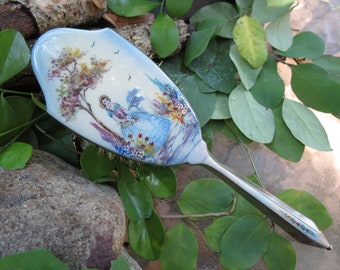May include: A silver hand mirror with a painted scene of a woman in a blue dress walking through a garden with flowers and trees. The mirror has a decorative handle with painted flowers.