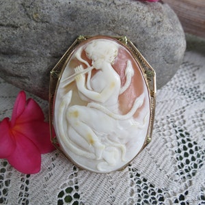May include: A cameo brooch featuring a woman playing a flute, surrounded by snakes, set in a gold tone frame.