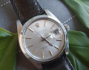 May include: A silver Rolex Oyster Perpetual Date wristwatch with a black leather band. The watch face has gold accents and the text "Rolex Oyster Perpetual Date Superlative Chronometer Officially Certified".