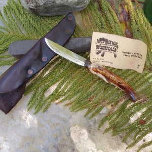 May include: A small, brown leather sheath with a single, copper-colored rivet holds a small,  knife with a brown antler handle. The knife blade is silver and has a slight curve. The knife is resting on a green leafy surface. A business card with the text "Mountain Man Originals" is visible in the background.