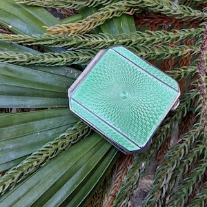 May include: A silver compact with a green enamel design. The design features a circular pattern with a concentric circle in the center.