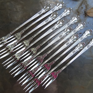 May include: A set of twelve silver-plated forks with ornate handles. The forks are arranged in a row, with the handles pointing towards the top of the image.