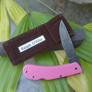 May include: A pink folding knife with a patterned blade and a brown suede case. The case has a white label with the text "Ralph D'Elia". The knife is open, revealing the blade. The knife is resting on a green leaf.