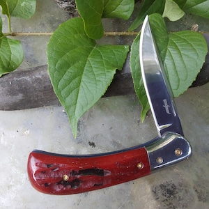 May include: A red and silver pocket knife with a wood handle and a stainless steel blade. The knife is open and the blade is sharp.