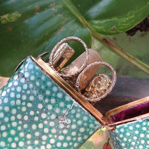 May include: A green and white spotted, hinged, rectangular box with a gold trim. The box is open and contains a gold thimble, a gold needle threader, and a gold needle case.