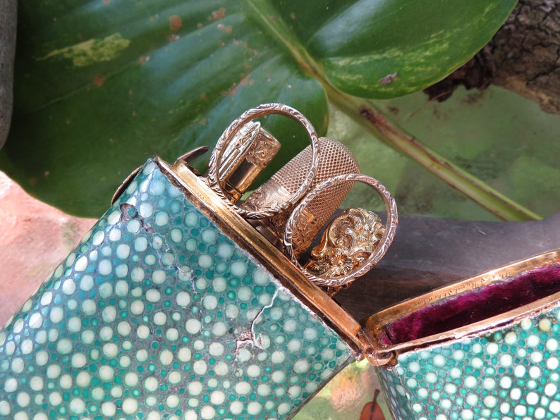 May include: A green and white spotted, hinged, rectangular box with a gold trim. The box is open and contains a gold thimble, a gold needle threader, and a gold needle case.