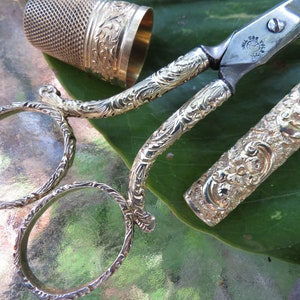 May include: A set of antique gold-toned scissors with a decorative floral design. The scissors are in a case with a matching design. The case is open and the scissors are partially extended.