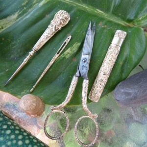 May include: A set of antique silver sewing tools on a green leaf. The set includes a thimble, a needle threader, a pair of scissors, and a needle case.