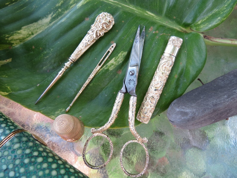 May include: A set of antique silver sewing tools on a green leaf. The set includes a thimble, a needle threader, a pair of scissors, and a needle case.
