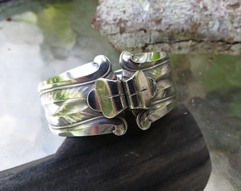 May include: A silver bracelet with a hinged clasp and a decorative design of leaves. The bracelet is made from a repurposed spoon.