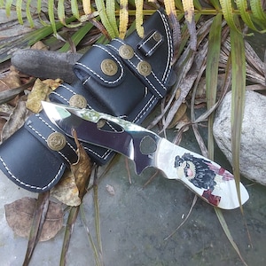 May include: A black leather sheath with white stitching holds a silver knife with a black and white cartoon animal design. The knife has a black handle and a silver blade.