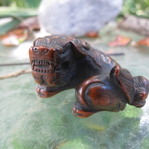 May include: A small, intricately carved wooden sculpture of a snarling lion with a fierce expression. The sculpture is made of dark brown wood and has a reddish-brown patina. The lion's eyes are wide and staring, and its teeth are bared in a menacing snarl.