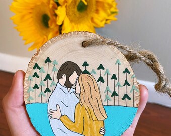 May include: A wooden ornament with a painted scene of a couple embracing in front of a forest and lake. The couple is painted in blue, yellow, and brown, while the forest is green and the lake is blue.
