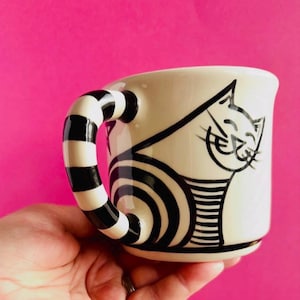 May include: A cream-coloured ceramic mug featuring a black line drawing of a cat with a smiling expression. The mug has a distinctive handle decorated with thick, horizontal black and white stripes that wrap around the entire handle structure.