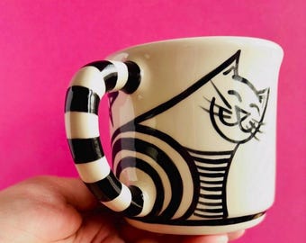 May include: A cream-colored ceramic mug featuring a black line drawing of a cat with a smiling expression. The mug has a distinctive handle decorated with thick, horizontal black and white stripes that wrap around the entire handle structure.