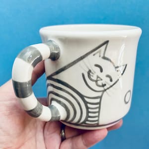 May include: A white ceramic mug with a grey and white striped handle. The mug features a smiling cat illustration in black and grey. The cat is wearing a striped shirt. The mug is held in a hand.