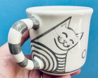 May include: A white ceramic mug with a gray and white striped handle. The mug features a smiling cat illustration in black and gray. The cat is wearing a striped shirt. The mug is held in a hand.