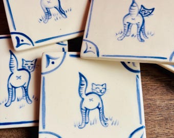 May include: Four white ceramic tiles with blue trim, each featuring a hand-painted illustration of a cat with its tail raised. The cats are depicted in a playful, cartoonish style.